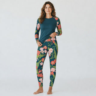 A woman modeling a long-sleeve rash guard top and matching floral swim leggings set. The set features a solid dark teal top with raglan sleeves and a dark teal base on the leggings covered in a colorful floral print with pink, red, and green flowers.