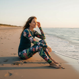 Woman in a colorful floral full coverage swimsuit sitting on a beach at sunset.