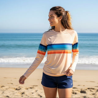 A woman stands on a sandy beach wearing a light peach long-sleeve athletic shirt with horizontal stripes in blue, teal, and orange, paired with navy shorts, with the ocean and blue sky in the background.